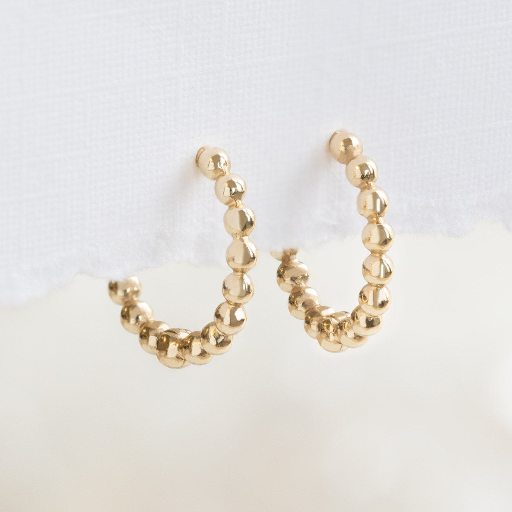 The Beaded Huggie Hoops in 14K Yellow Gold Hanging on Textured White Paper