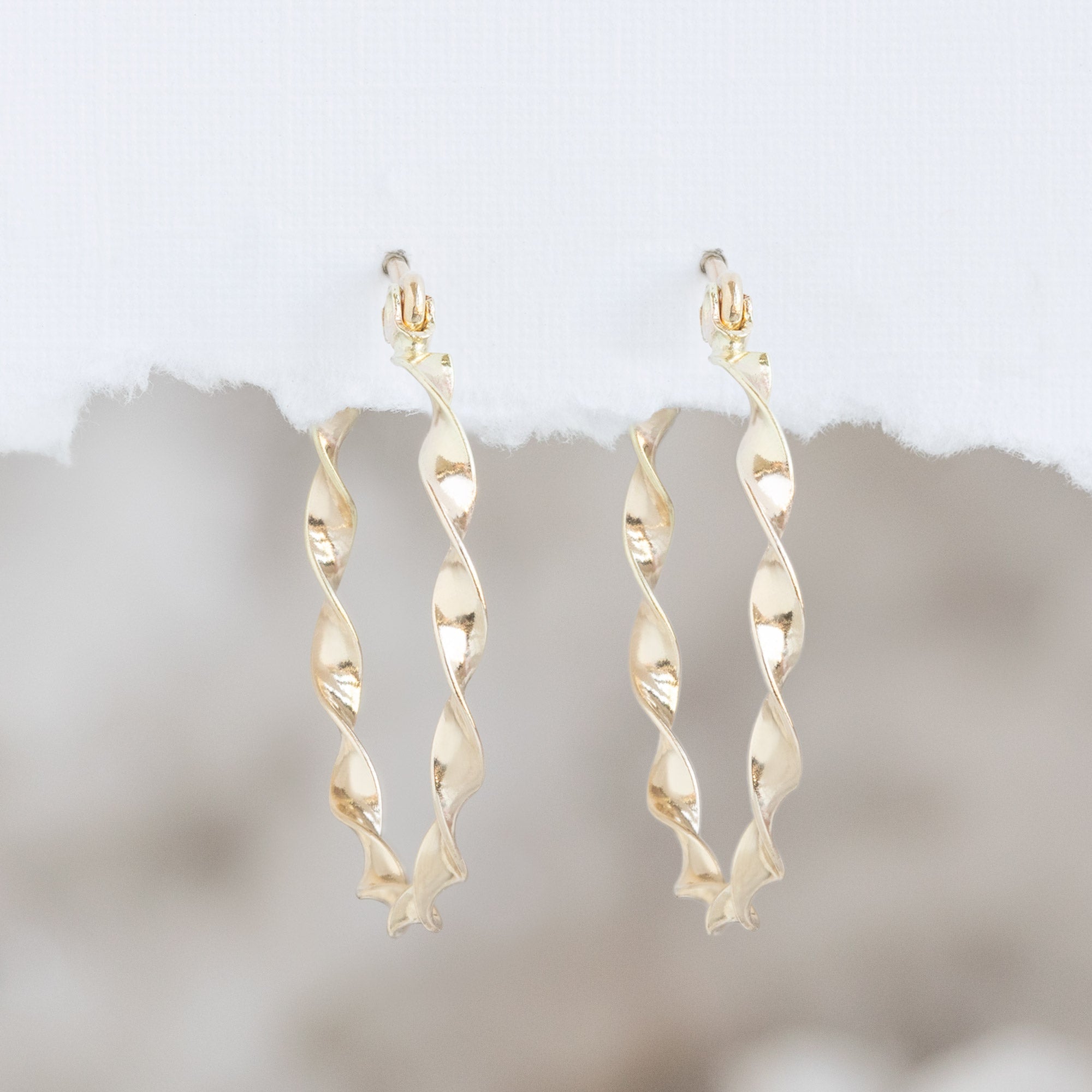 The Ribbon Hoops in 14K Yellow Gold hanging on textured white paper