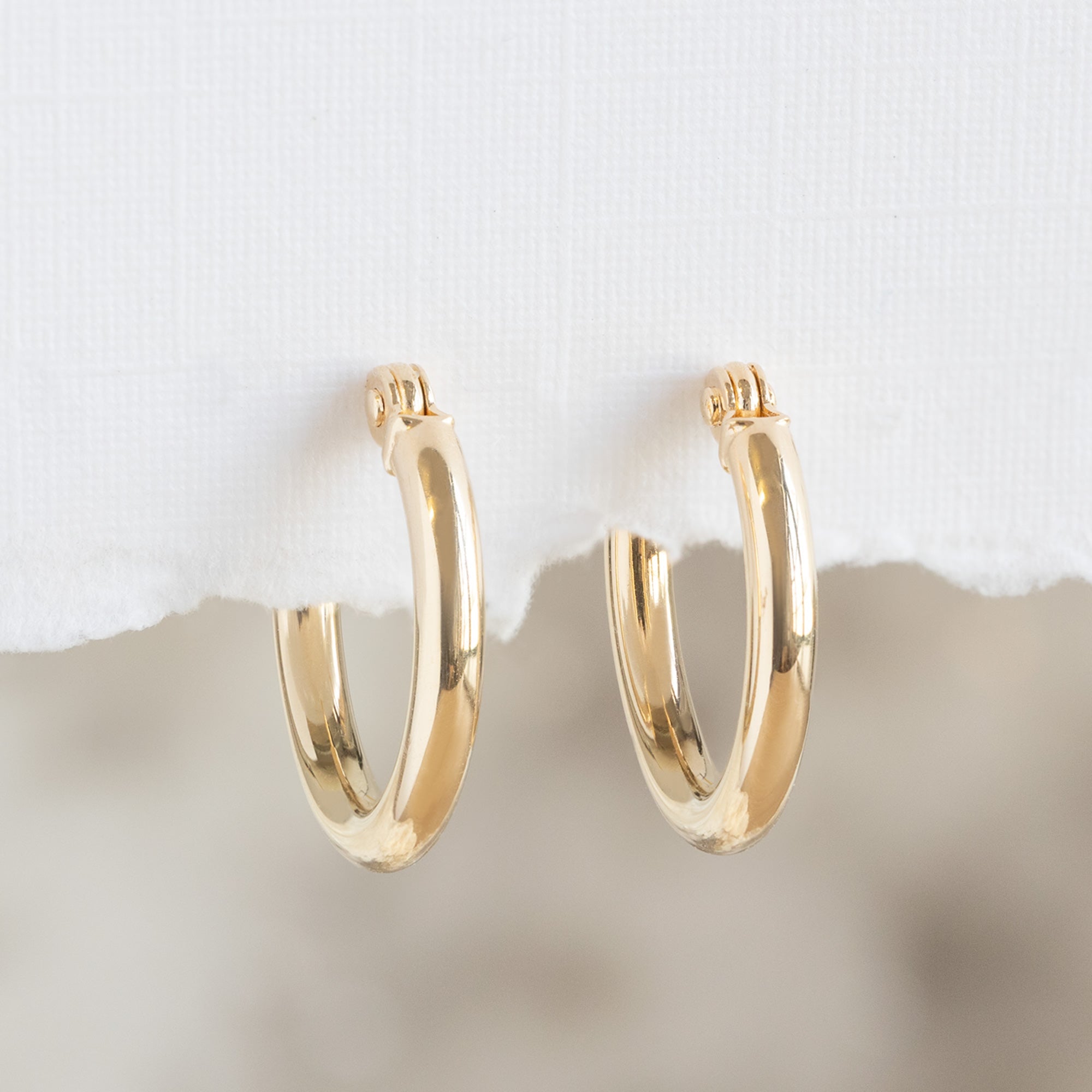 The Charm Hoop Earrings in 14K Yellow Gold hanging on white textured paper