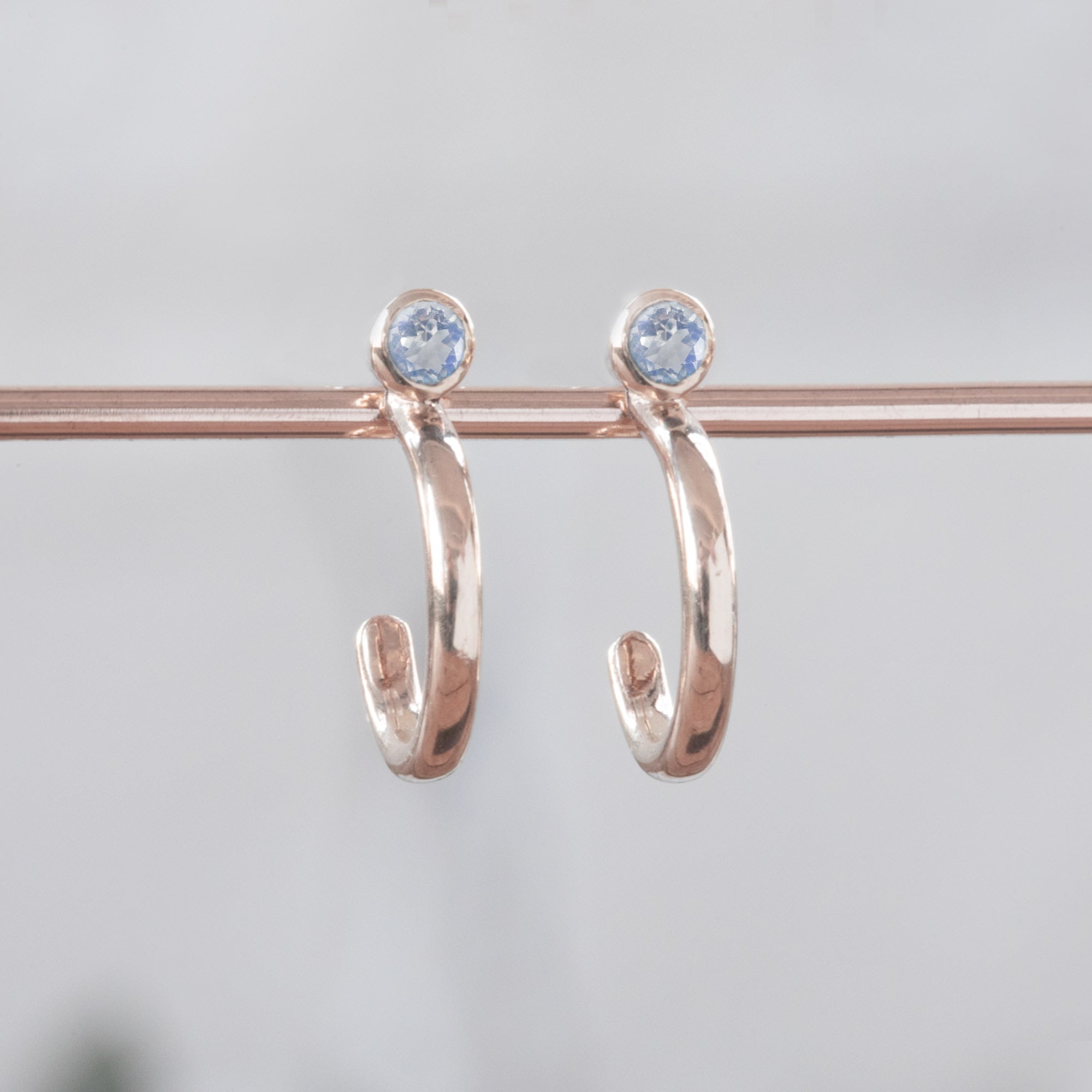 Custom Moonstone Birthstone Hoop Earrings | 14K Rose Gold. Displayed on holding bar with white blurred background.