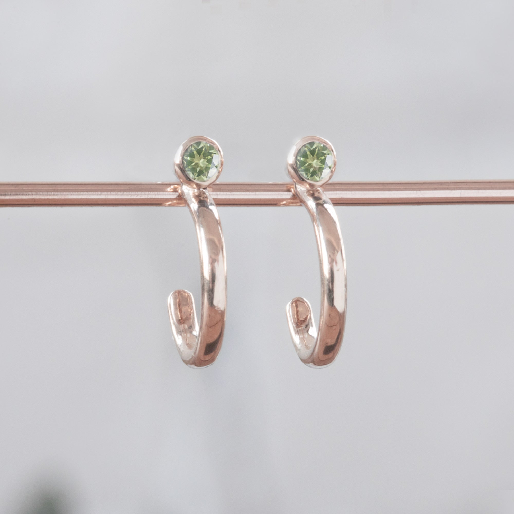 Custom Peridot Birthstone Hoop Earrings | 14K Rose Gold. Displayed on holding bar with white blurred background.