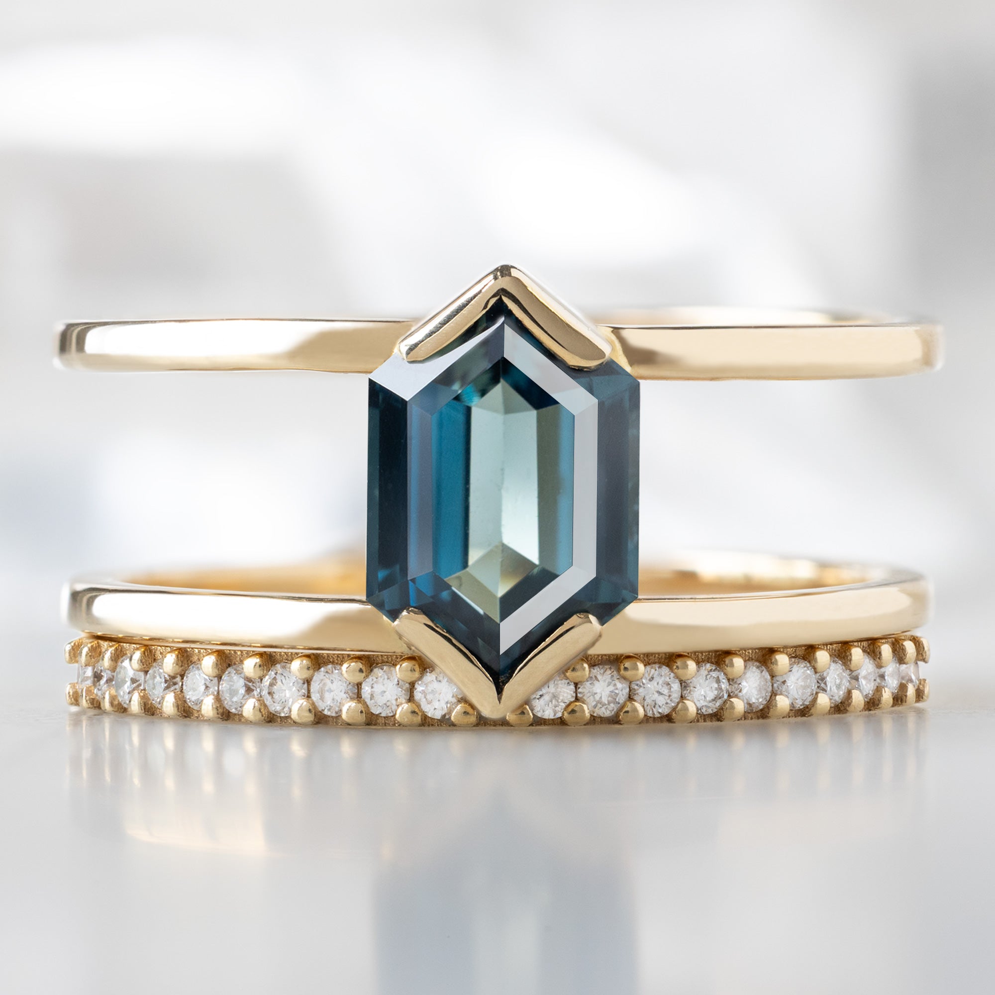 The Poppy Ring with a 1.24ct Hexagon Blue Sapphire in 14K Yellow Gold Stacked with the White Diamond Pavé Wedding Band on White Marble