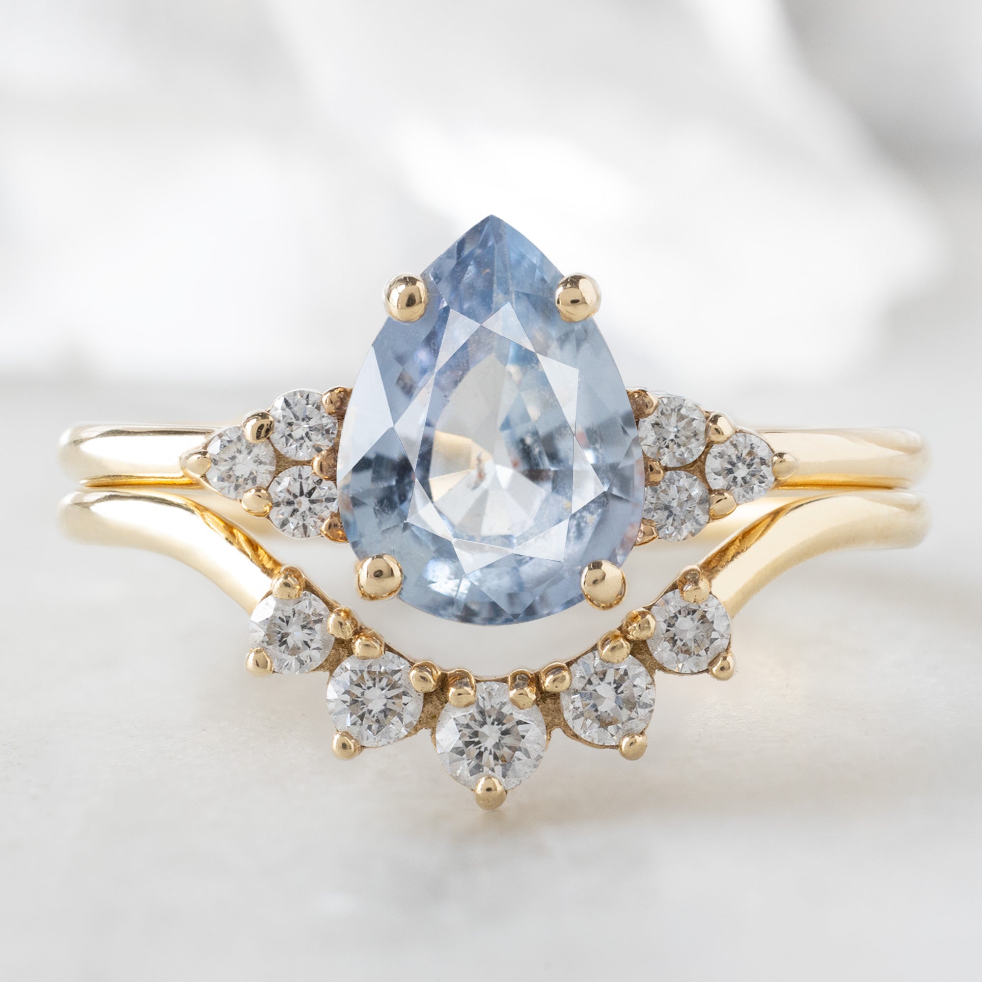 The Ivy Ring with a 1.5ct Pear Cut Sapphire in 14K Yellow Gold Stacked with the Round White Diamond Sunburst Stacking Band on White Marble