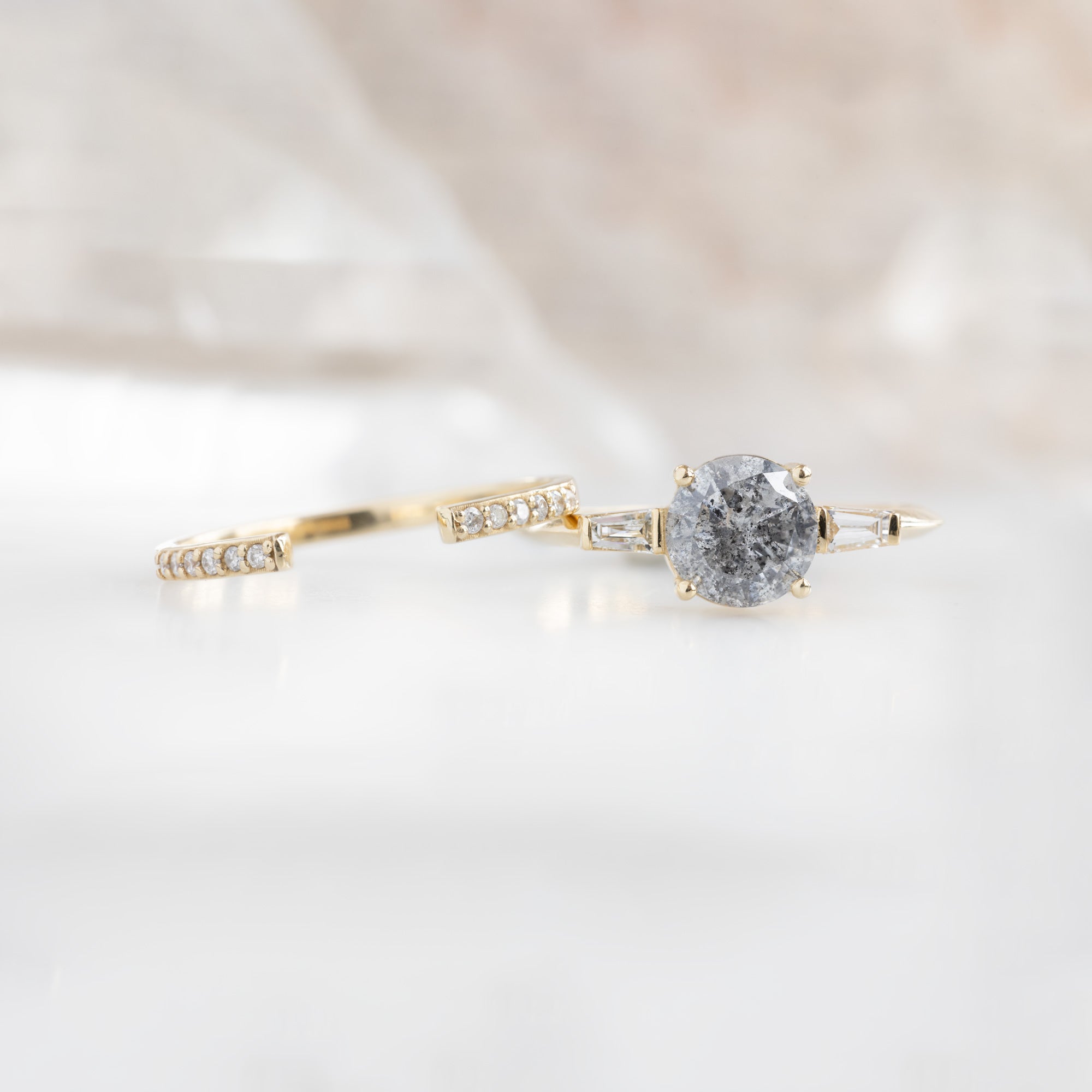 The Ash Ring | 1.22 carat  Round Salt and Pepper Diamond in 14K Yellow Gold with white blurred background. displayed next to the open cuff flush pavé wedding band.