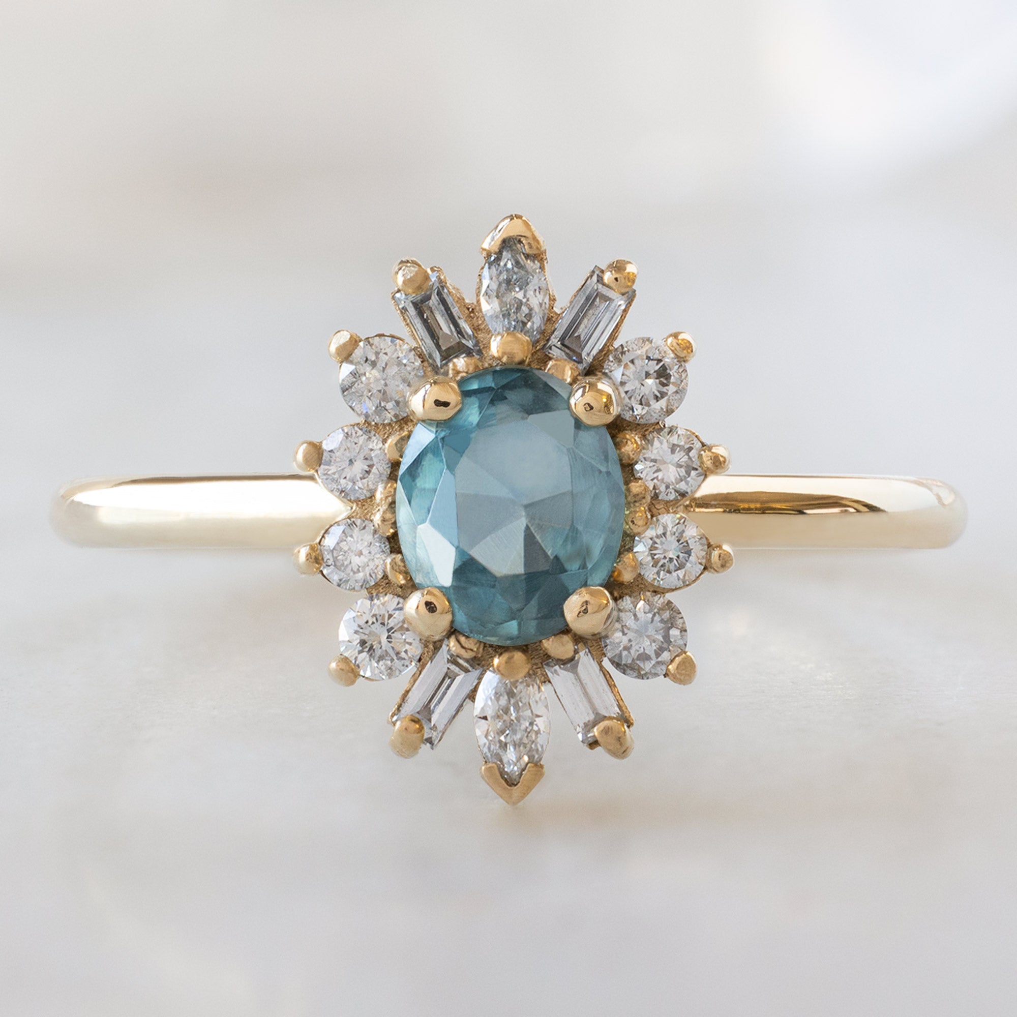 The Camellia Ring with an Oval Cut Blue Sapphire in 14K Yellow Gold on White Marble