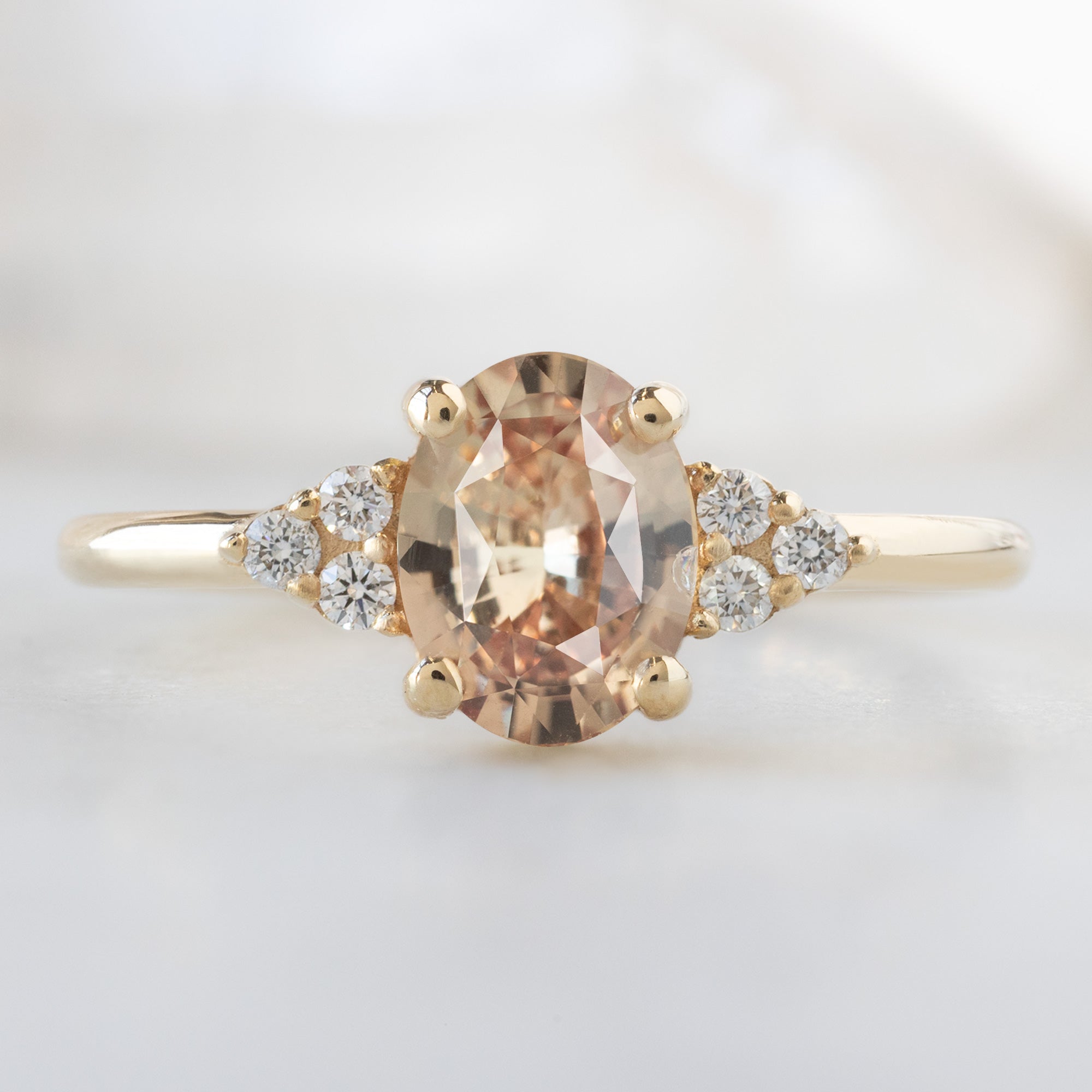 The Ivy Ring with a 1.13ct Oval Peach Sapphire in 14K Yellow Gold on White Marble Surface