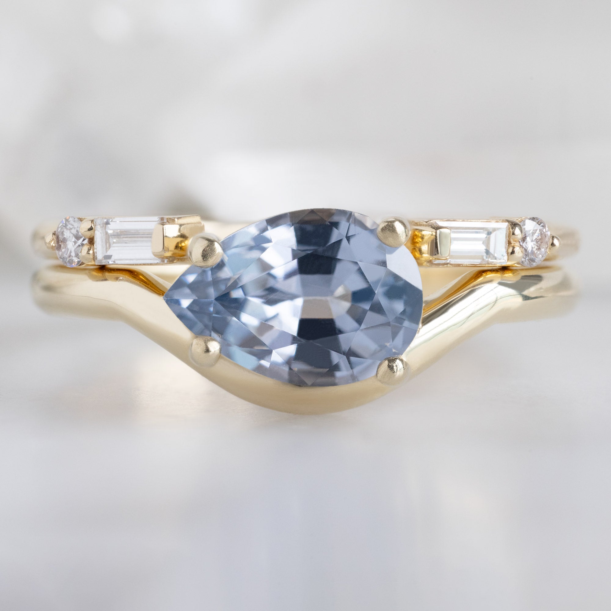 The Wave Ring with a 1.29ct Pear Lavender Spinel in 14k Yellow Gold Stacked with the Lux Open Cuff Baguette Stacking Band on White Marble Surface