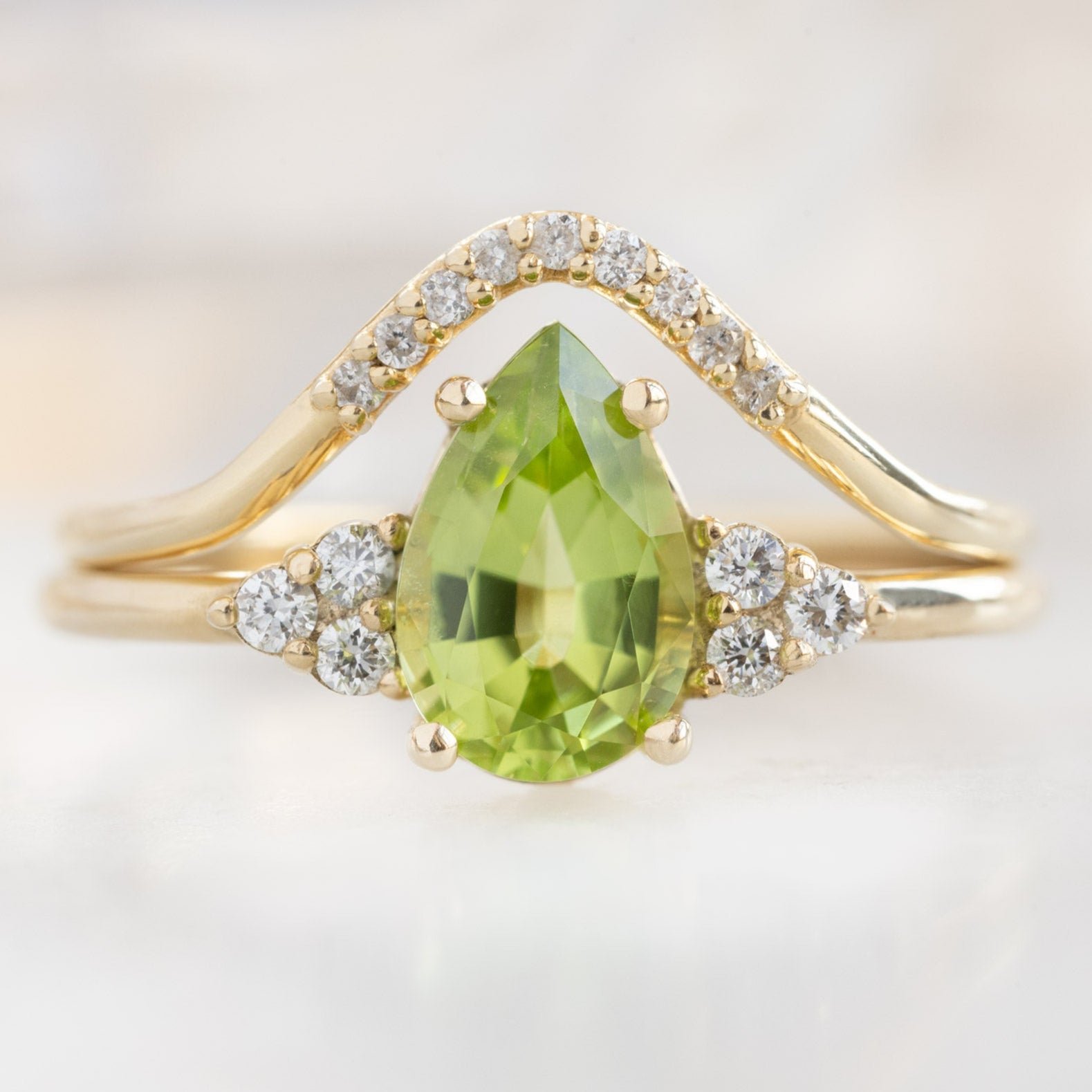 The Ivy Ring with a .89ct Pear Peridot in 14K Yellow Gold Stacked with the White Pavé Diamond Peak Stacking Band on White Marble Surface