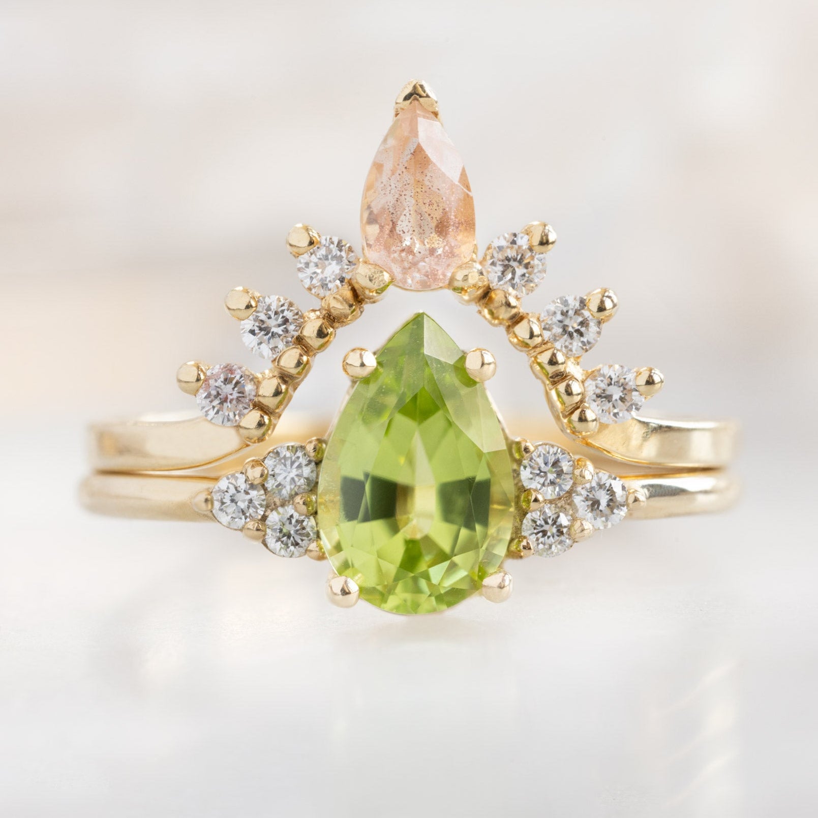 The Ivy Ring with a .89ct Pear Peridot in 14K Yellow Gold Stacked with the Sunstone and White Diamond Sunburst Stacking Band on White Marble Surface