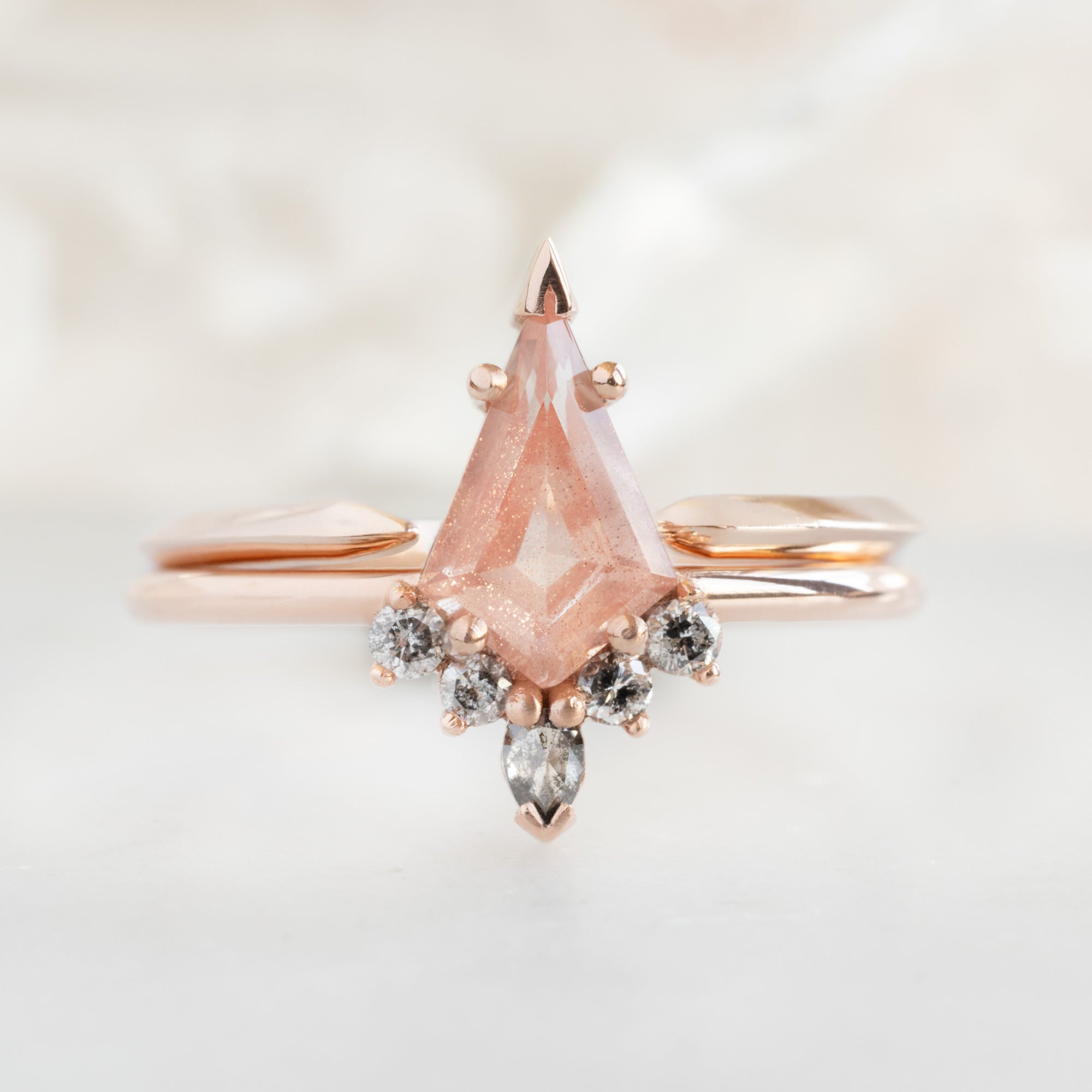 The Aster Ring | 0.79 carat Kite Sunstone in 14K Rose Gold with white blurred background. Displayed next to the open cuff knife edge wedding band.