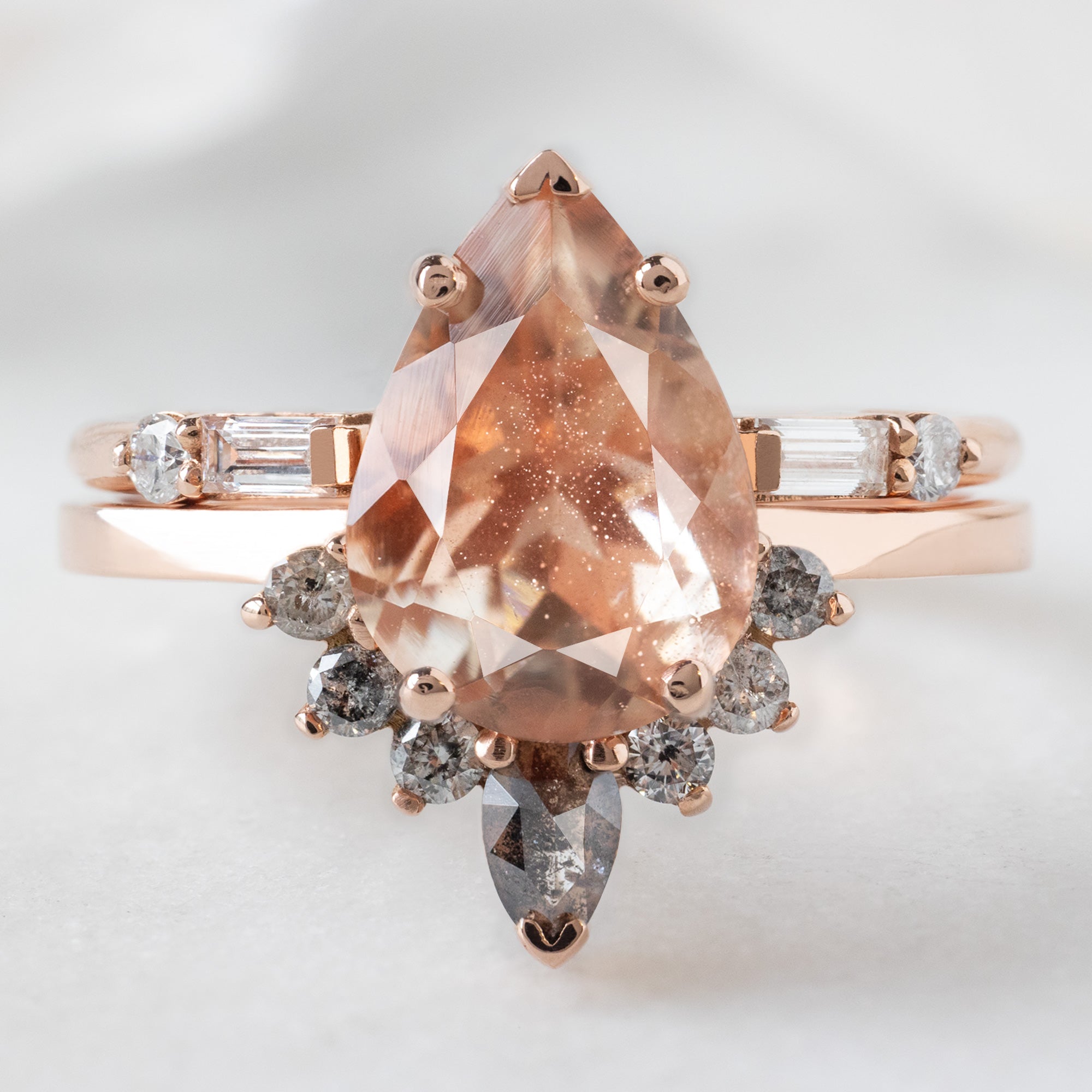 The Aster Ring with a 1.96ct Pear Sunstone in 14K Rose Gold  Stacked with the Luxe Open Cuff White Baguette Diamond Stacking Band on White Marble Surface