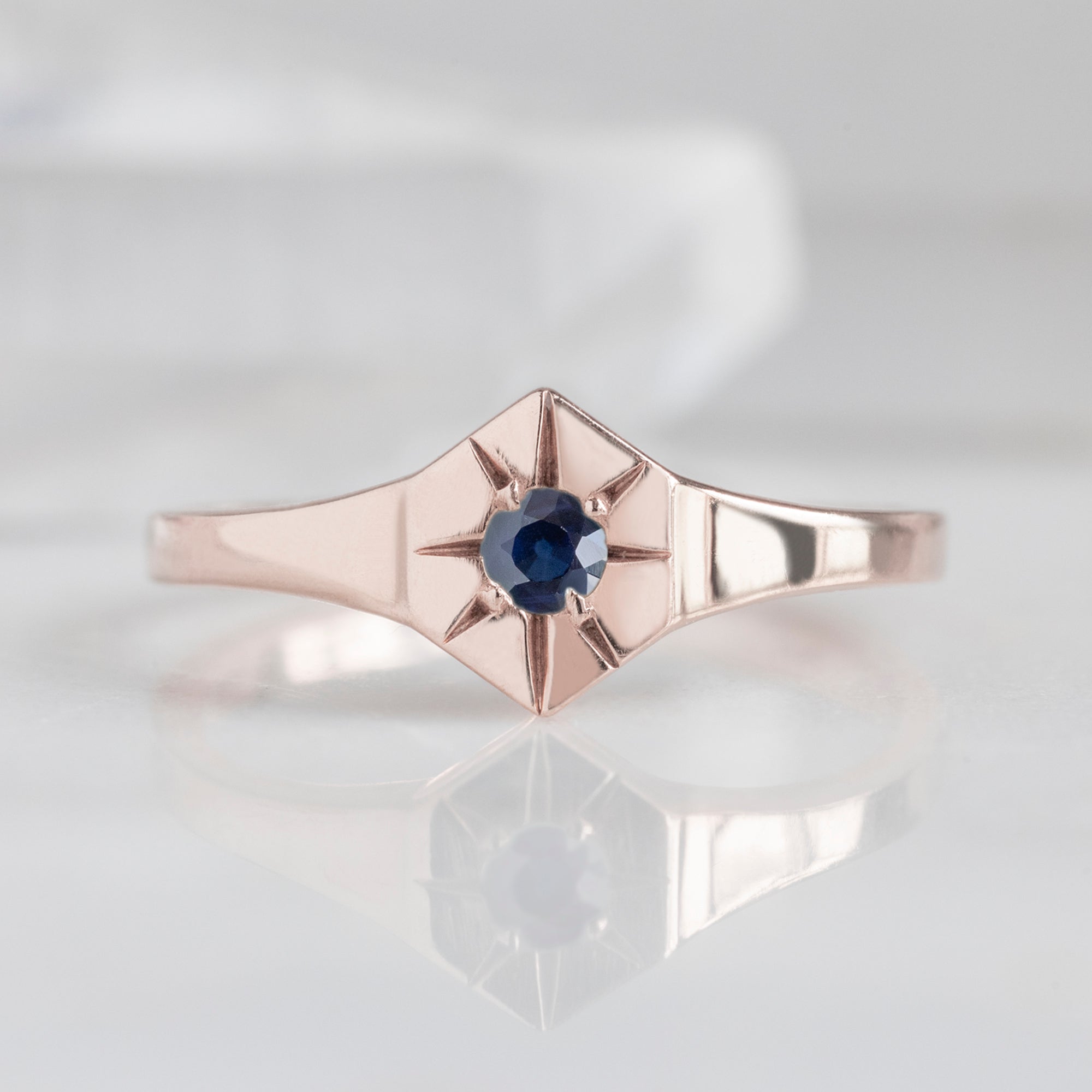 Custom Blue Sapphire Birthstone Signet Ring | 10K Rose Gold displayed on marble surface with white blurred background.