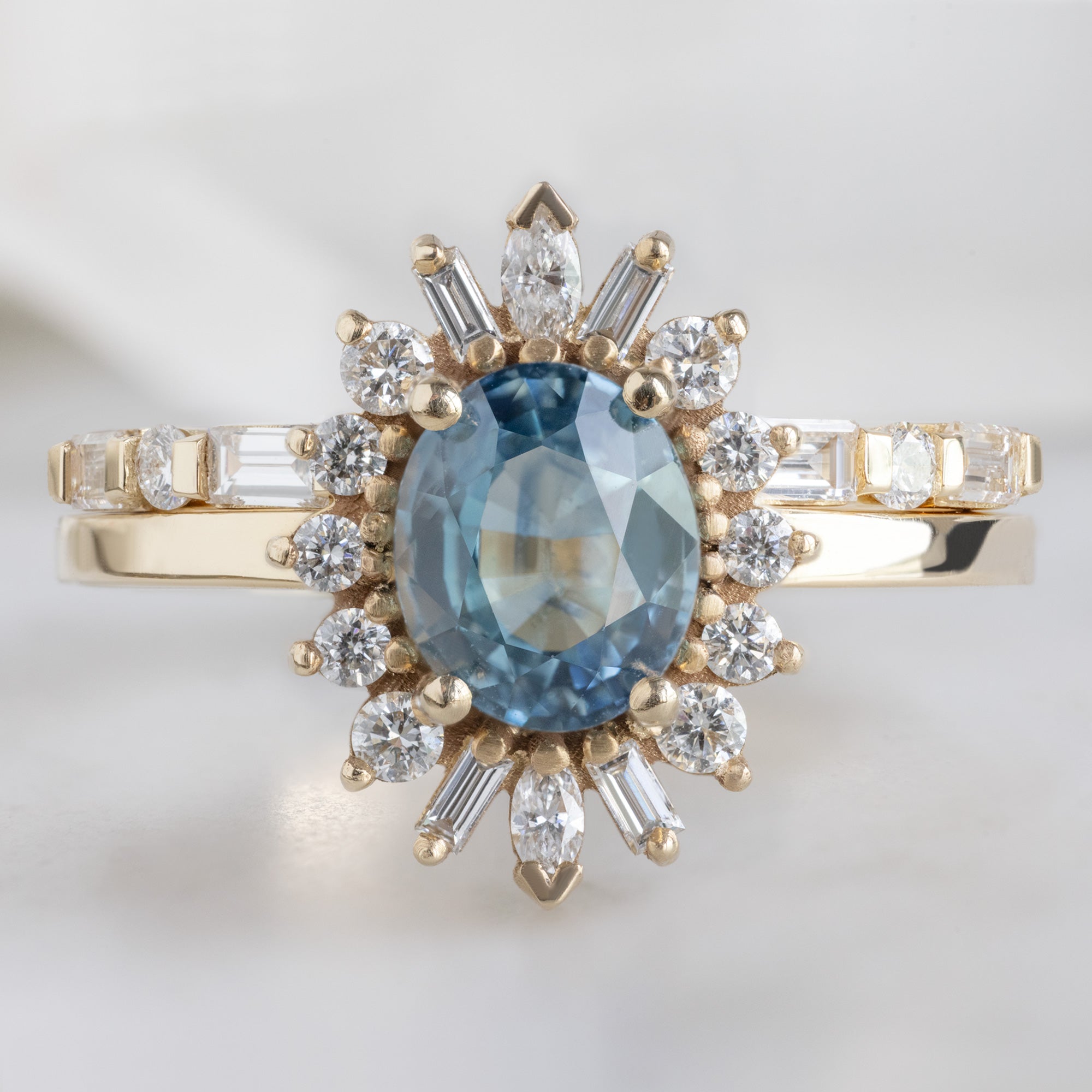 The Luxe Baguette White Diamond Eternity Band in 14K Yellow Gold stacked with oval sapphire gemstone engagement ring on white marble surface