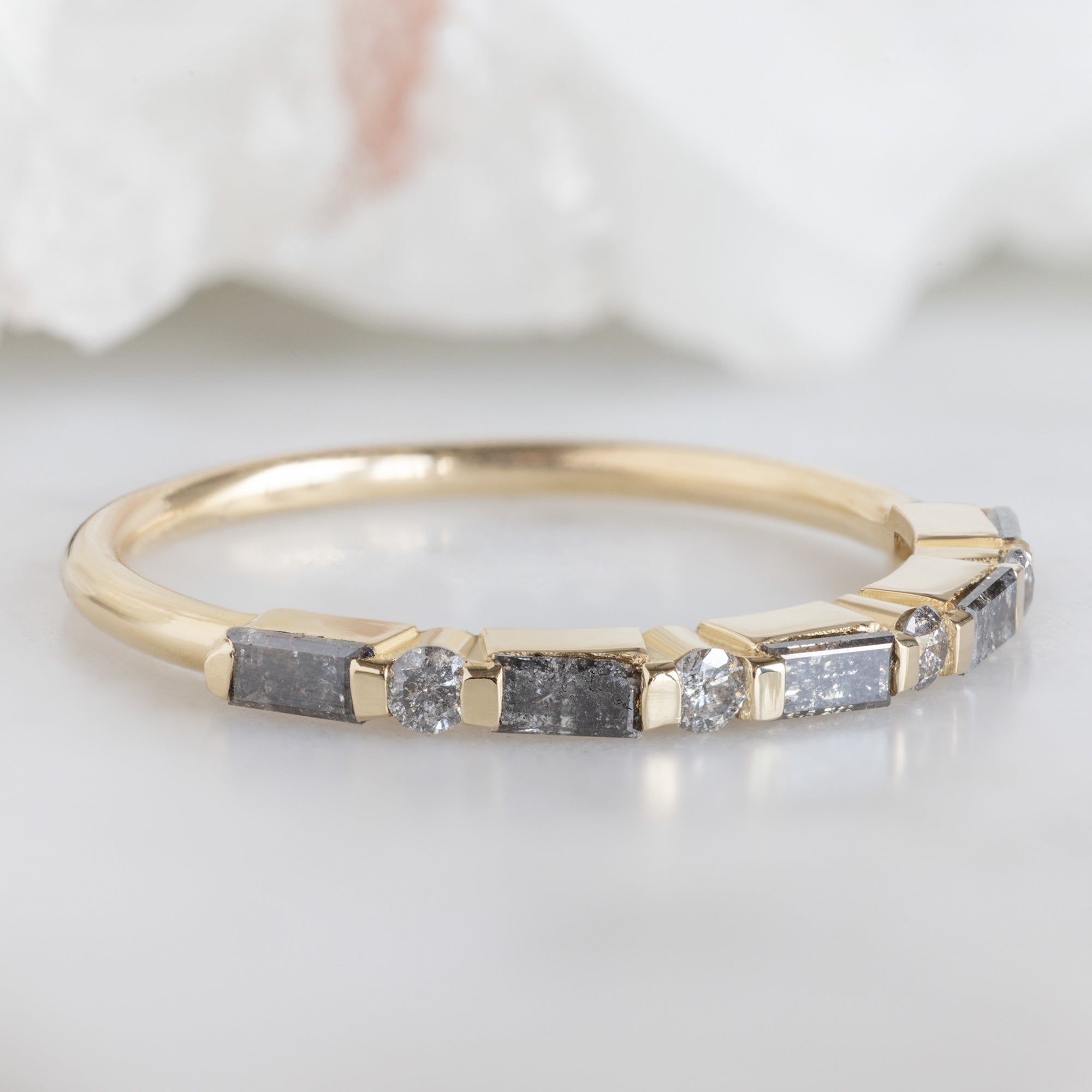 The Luxe Baguette Salt and Pepper Diamond Eternity Band in 14K Yellow Gold on white marble surface