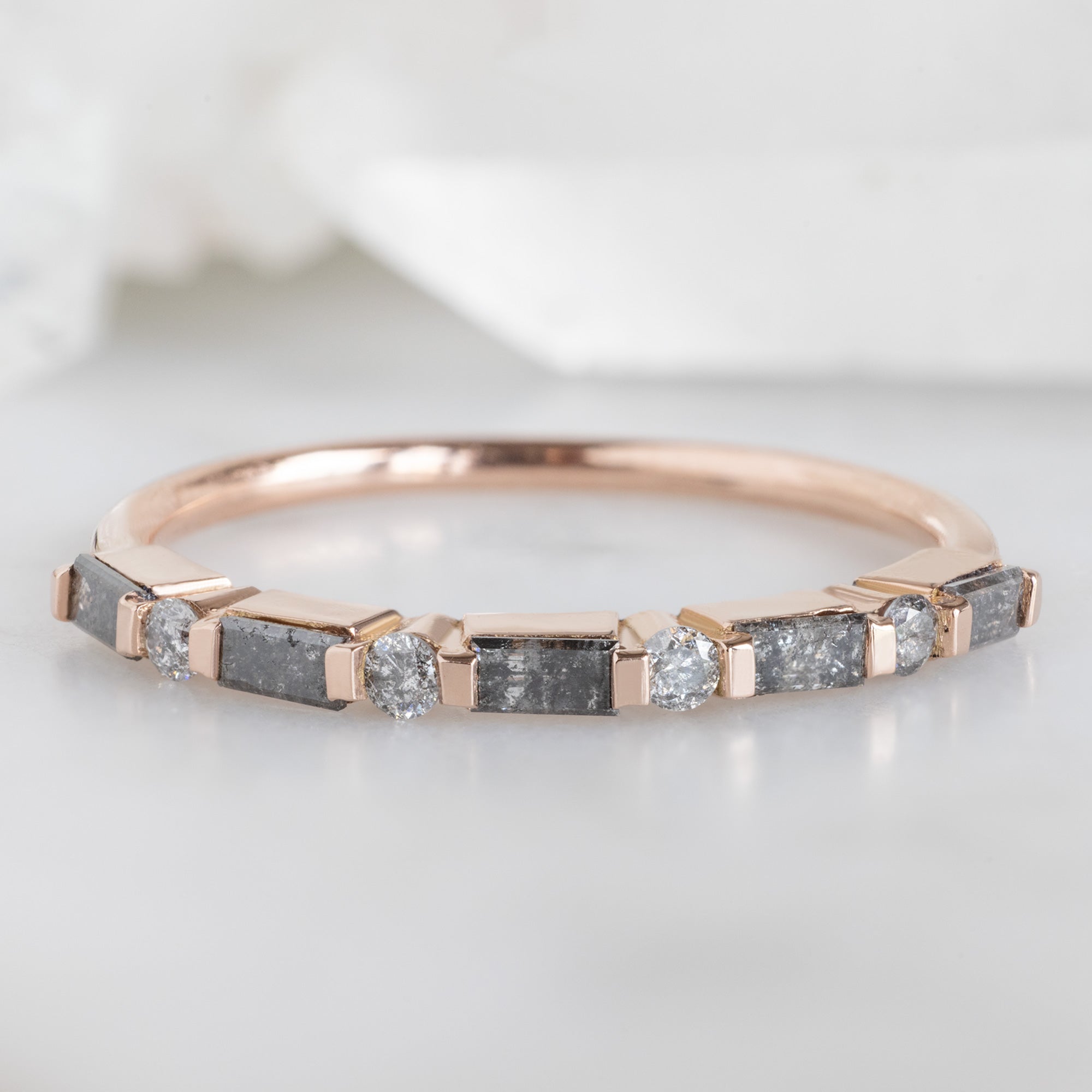 The Luxe Baguette Salt and Pepper Diamond Eternity Band in 14K Rose Gold on White Marble Surface