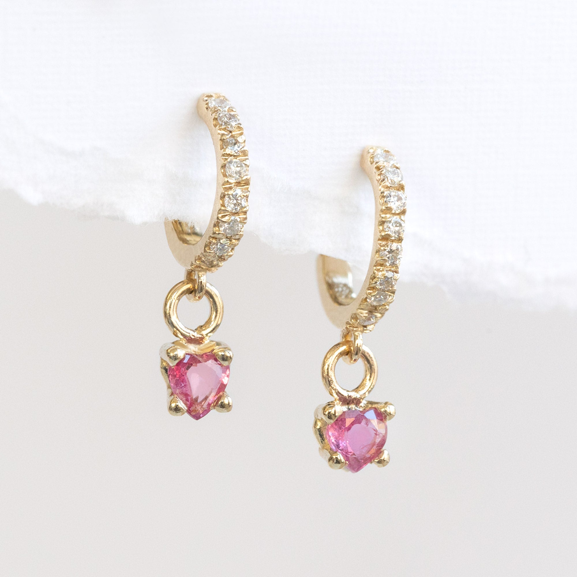 The Ruby Heart + Diamond Huggie Studs in 14K Yellow Gold hanging on White Textured Paper