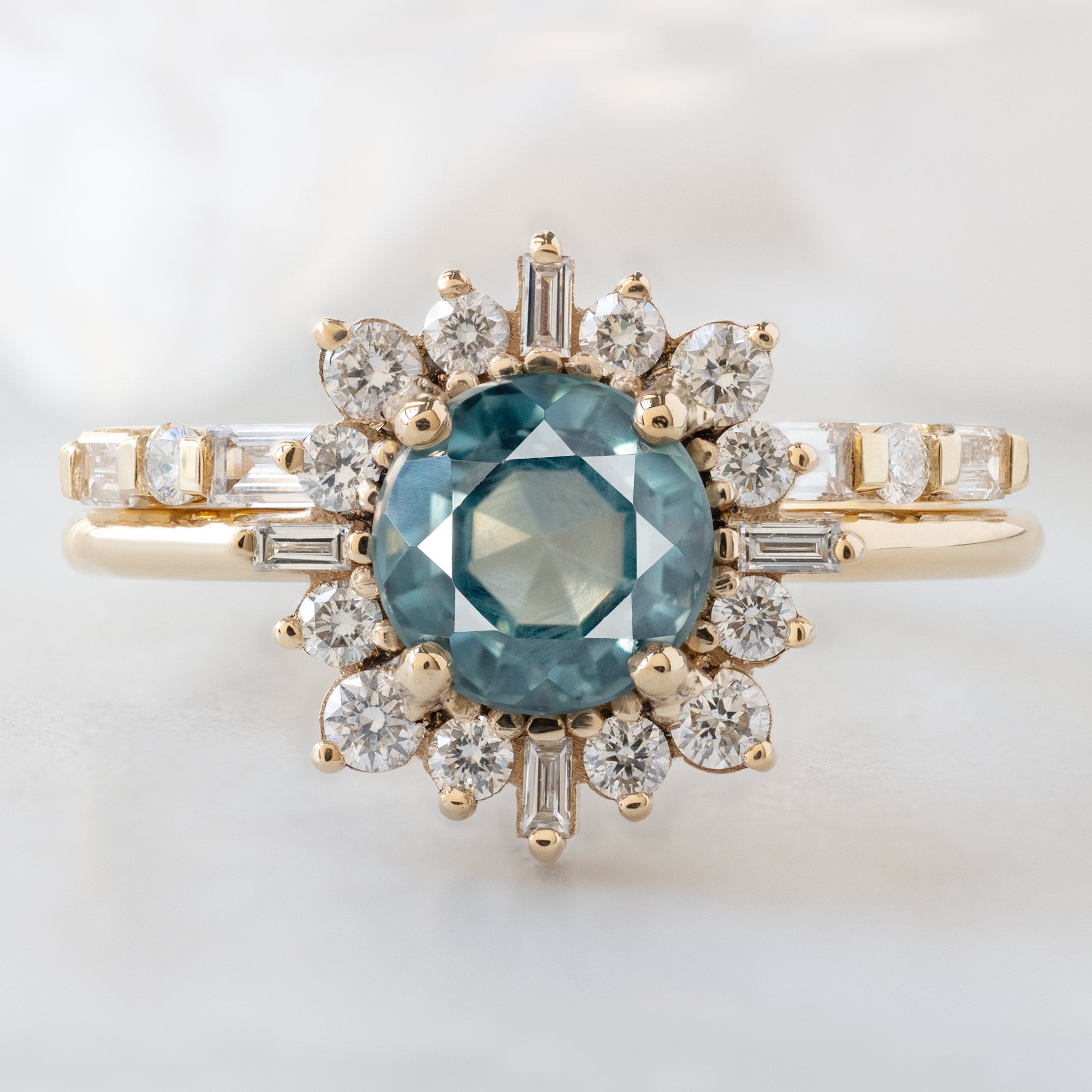 The Compass Ring with a 1.66ct Round Blue Sapphire in 14K Yellow Gold Stacked with the Luxe Open Cuff Baguette White Diamond Stacking Ring on White Marble Surface