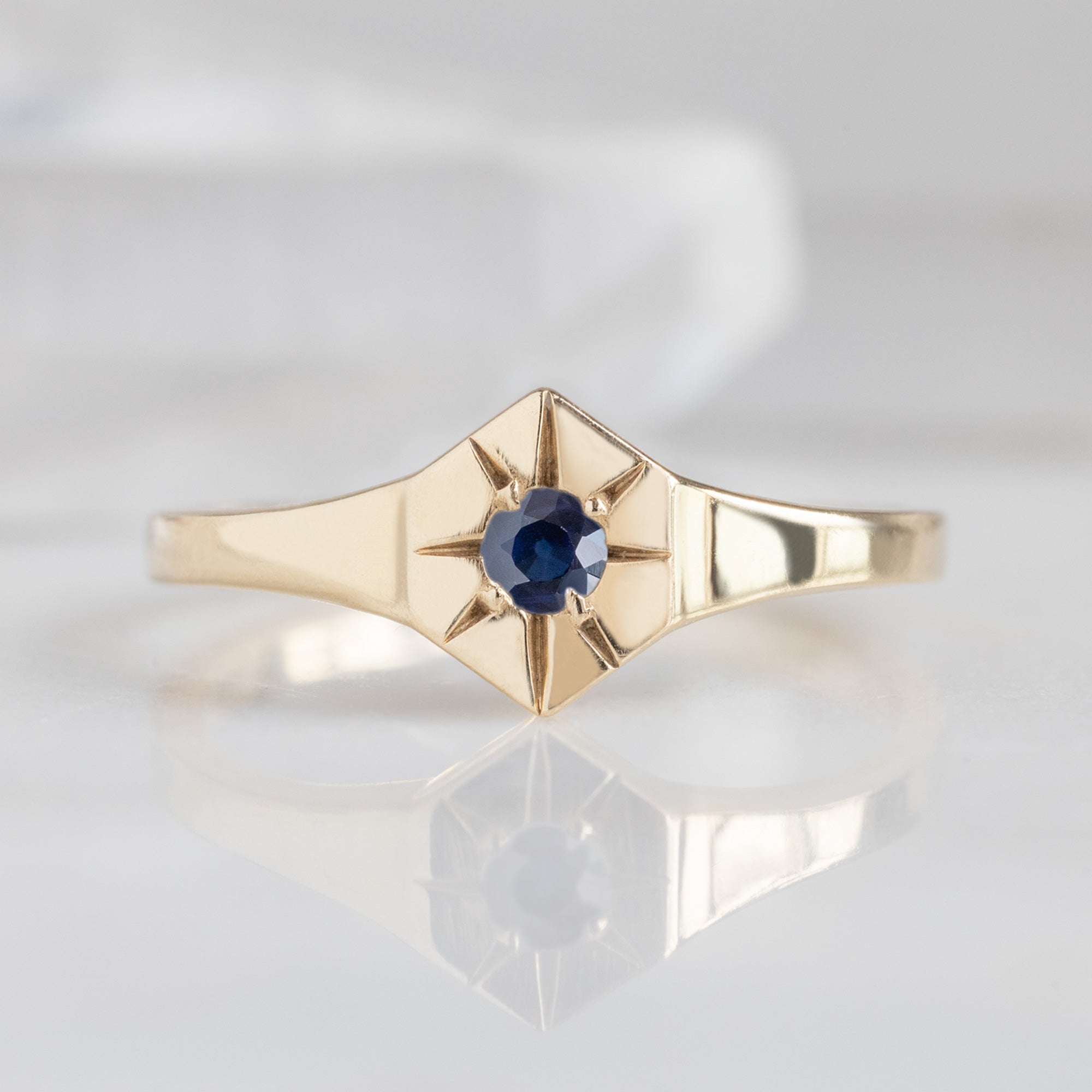 Custom Blue Sapphire Birthstone Signet Ring | 10K Yellow Gold displayed on marble surface with white blurred background. 